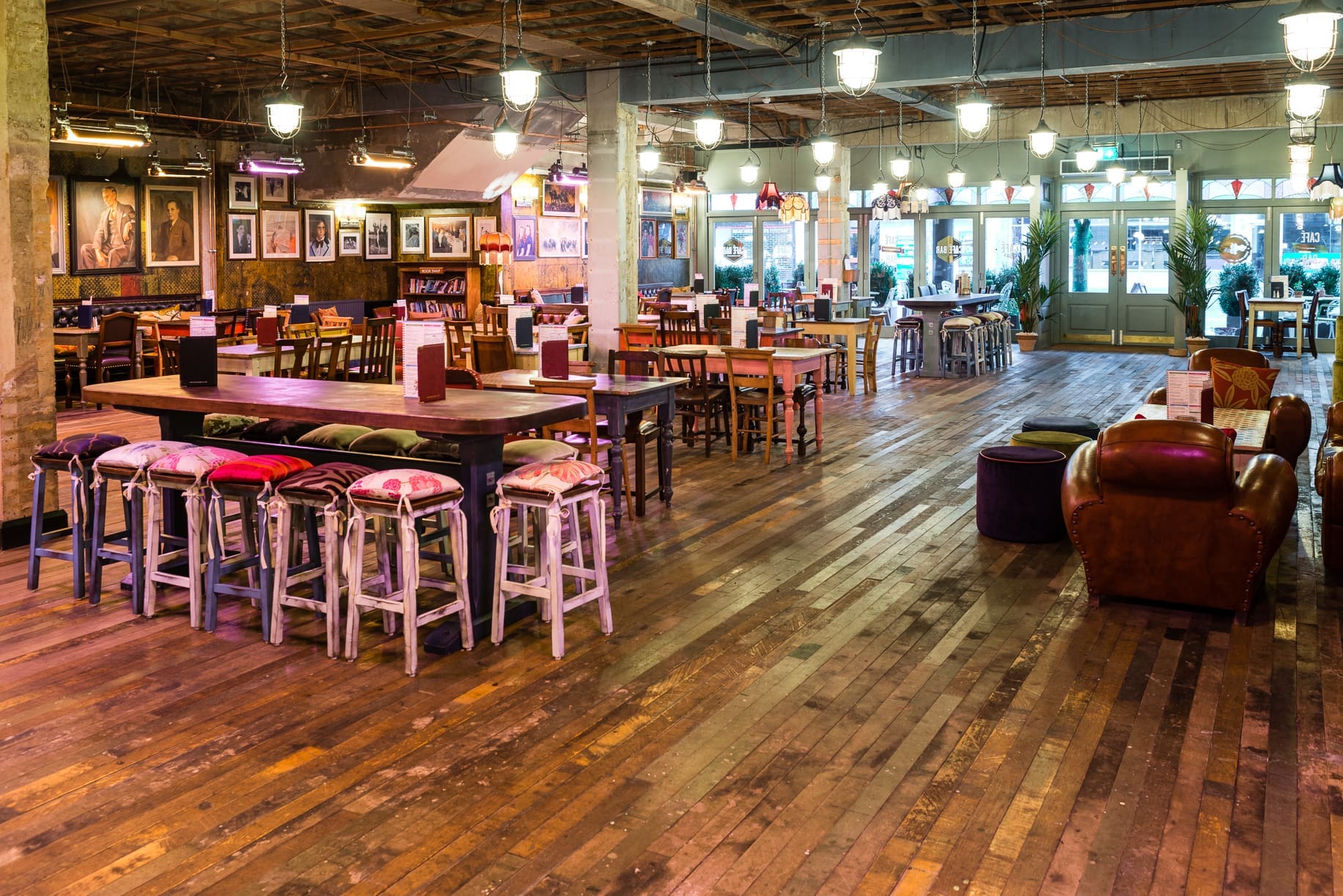 A spacious, rustic-style cafe or bar near Alcampo with wooden floors, mismatched tables and chairs, colorful cushions, cozy armchairs, wall art, and large windows letting in natural light.