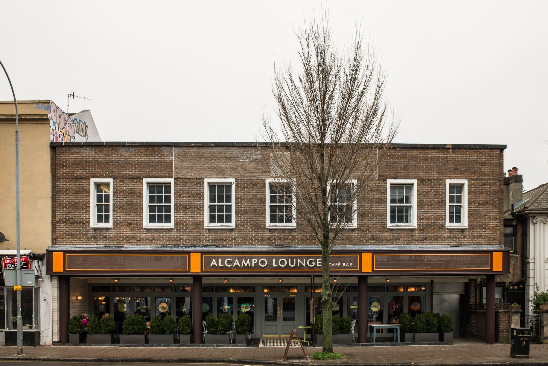 A two-story brick building with large windows and a sign reading "Alcampo Lounge Café Bar" above the entrance. The Alcampo features glass doors, plants, and a leafless tree in front, inviting guests to relax inside.