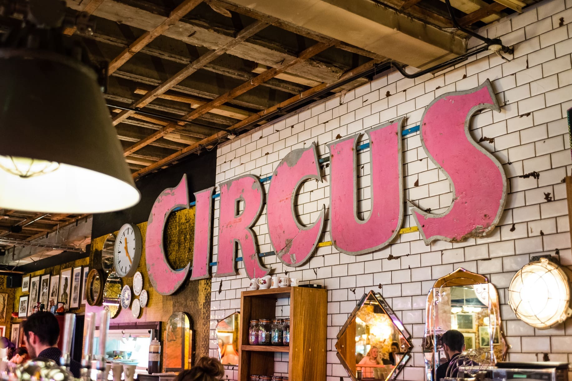 A large, pink "CIRCUS" sign is mounted on a white tiled wall in a lively, eclectic indoor space—reminiscent of Alcampo—decorated with vintage mirrors, clocks, and shelves.