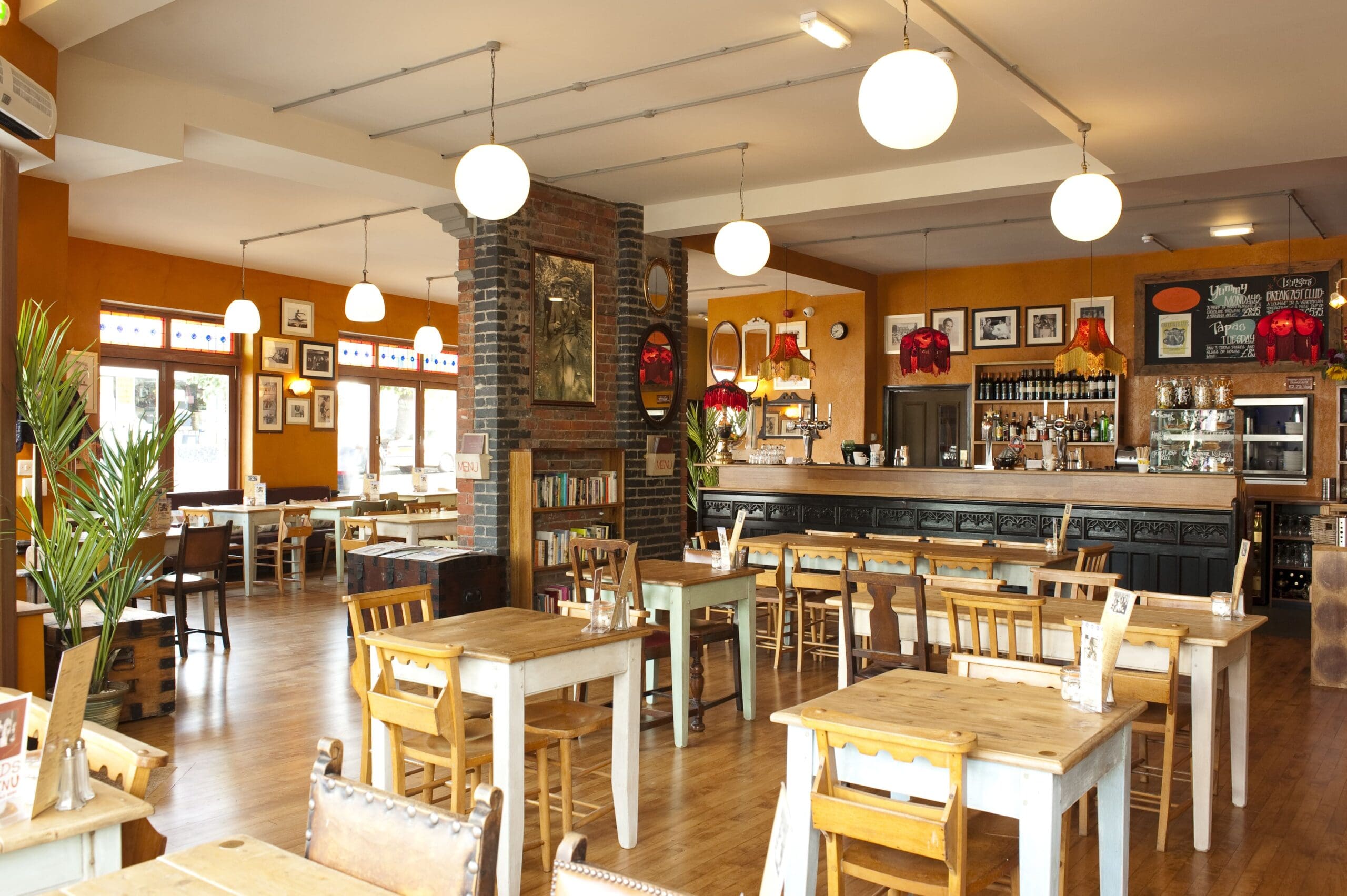 A cozy, warmly lit café named Santo features wooden tables and chairs, a bar counter at the back, framed pictures on orange walls, hanging globe lights, plants, and large windows letting in natural light.