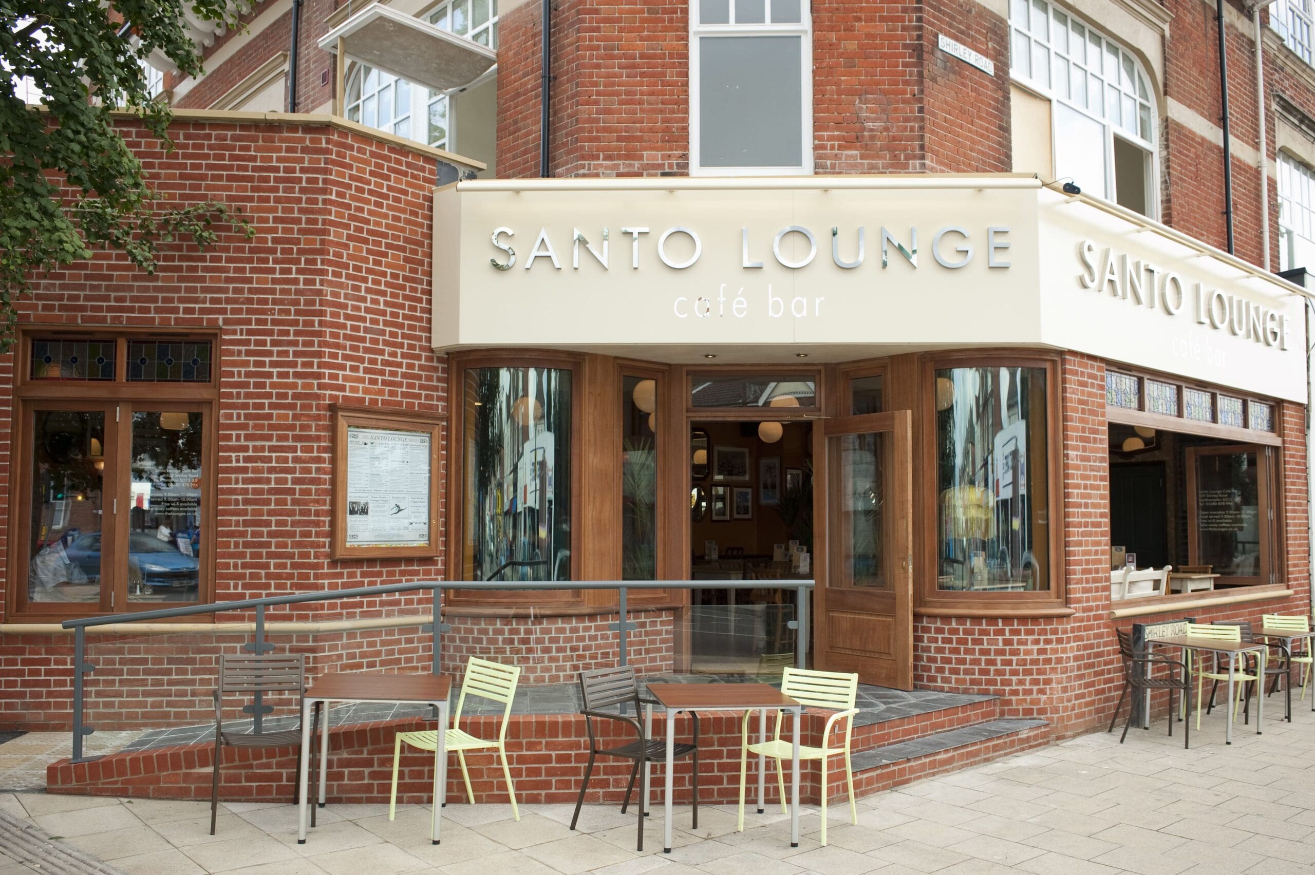 Exterior view of Santo Lounge café bar with large windows, wooden doors, colorful outdoor seating, and a brick facade showcasing the lively Santo atmosphere on a street corner.
