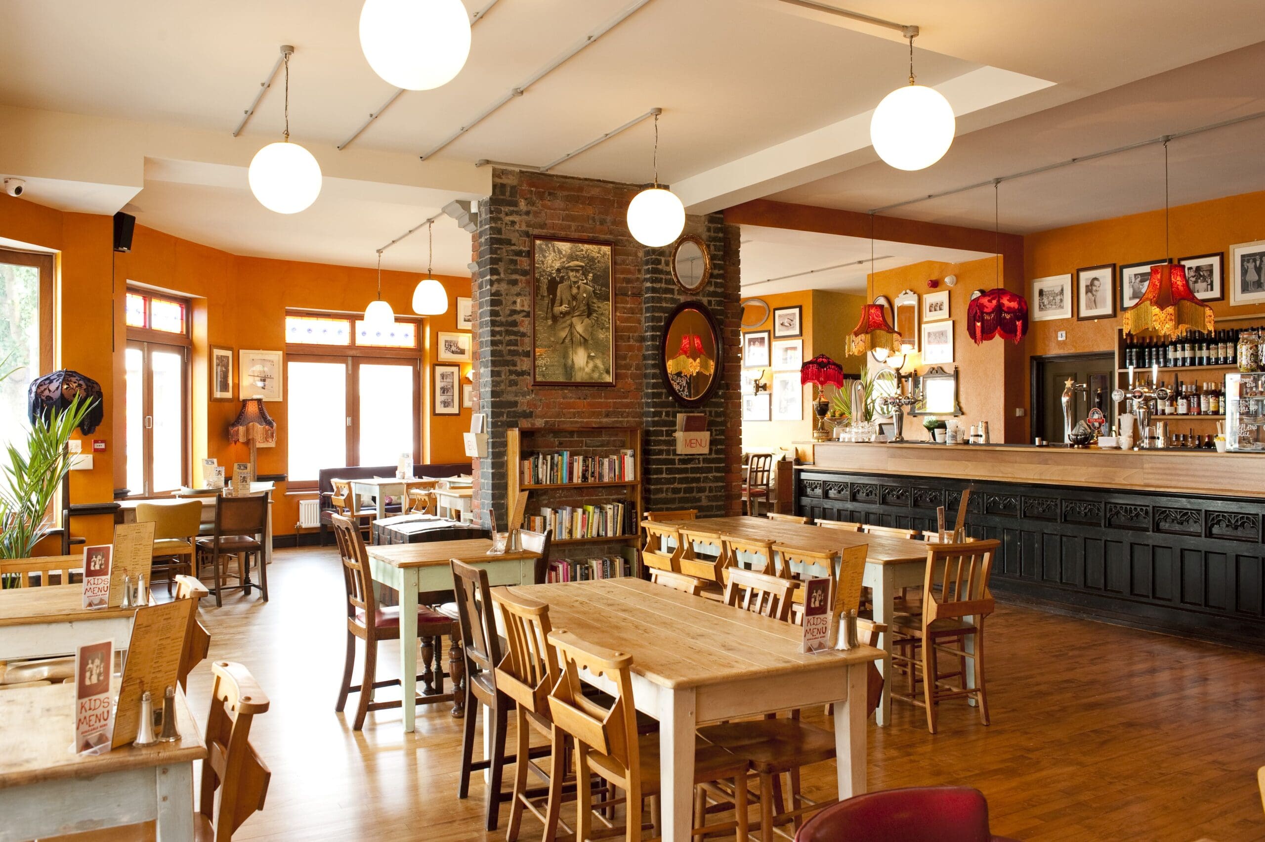 A cozy Santo cafe with wooden tables and chairs, warm lighting from round ceiling lamps, a bookshelf, framed pictures on orange-patterned walls, and a long bar counter on the right.