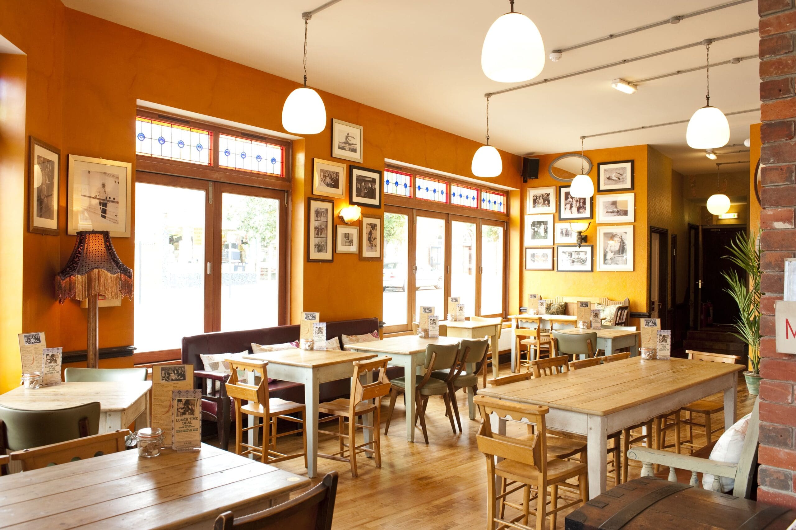 A cozy, well-lit café in Santo with wooden tables and chairs, large windows, framed pictures on orange walls, and hanging pendant lights creating a warm, inviting atmosphere.