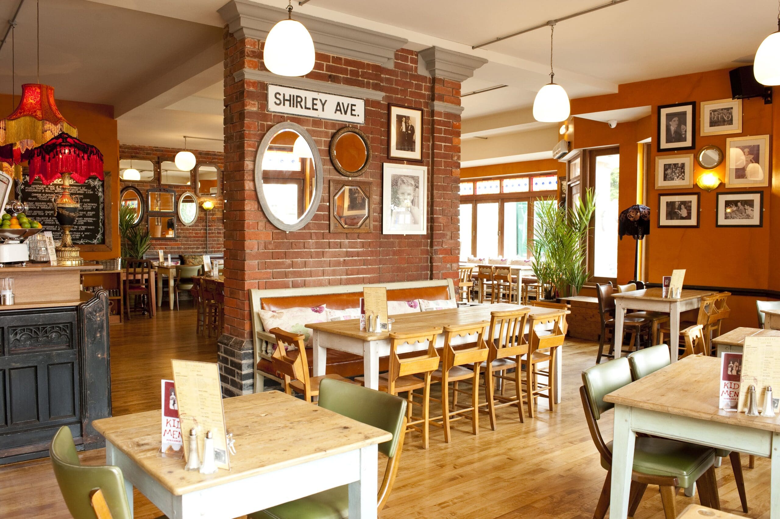 A cozy, brightly lit café with wooden tables and chairs, brick columns, and framed photos and mirrors on the walls—Santo-inspired touches and decorative plants add a lively vibe. A "SHIRLEY AVE." sign and large windows invite in plenty of natural light.