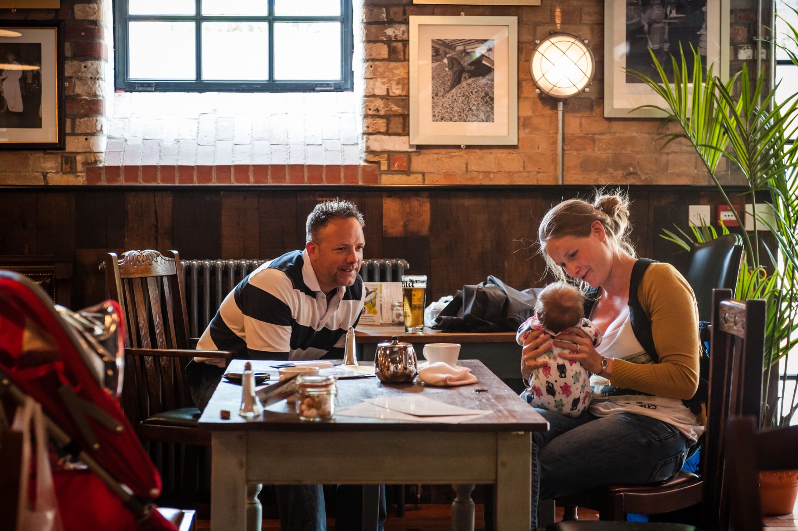 A man and woman sit at a rustic café table, smiling and interacting with a baby. The woman holds the baby on her lap while the man leans forward. Warm Vivo tones fill the cozy café, with wooden decor, framed photos, and soft lighting.