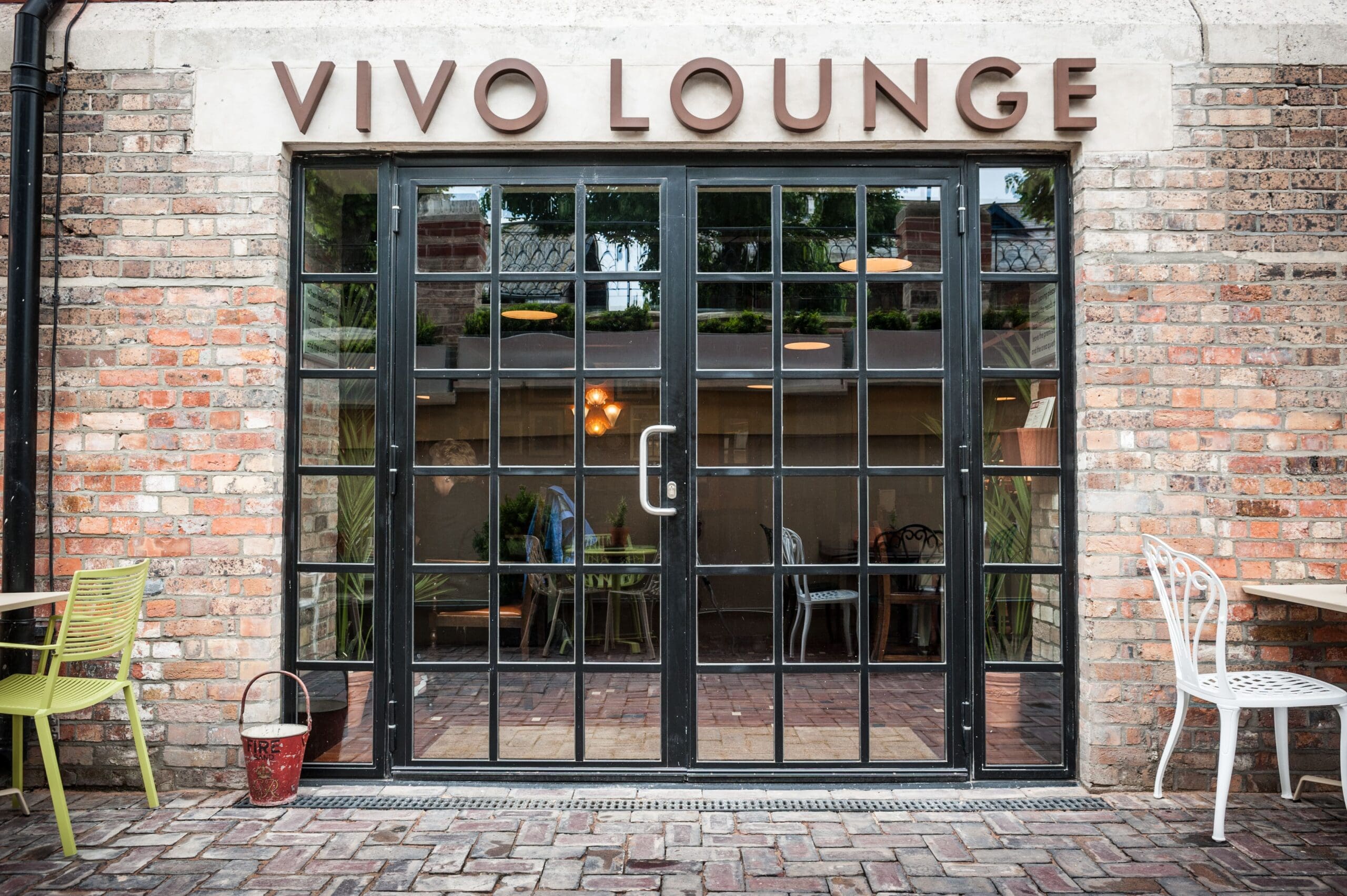 Exterior view of Vivo Lounge, featuring large glass doors with a metal grid frame, brick walls, and inviting outdoor seating with colorful chairs and tables arranged on the lively Vivo brick patio.