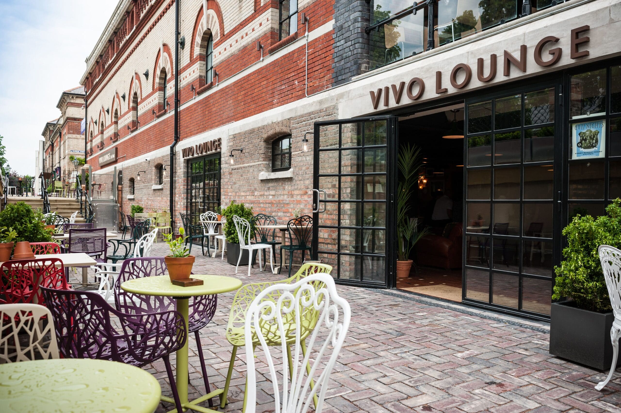The outdoor seating area of Vivo features colorful metal chairs and tables on a brick patio, set beside a large building with glass doors, lush potted plants, and vibrant red and tan brickwork.