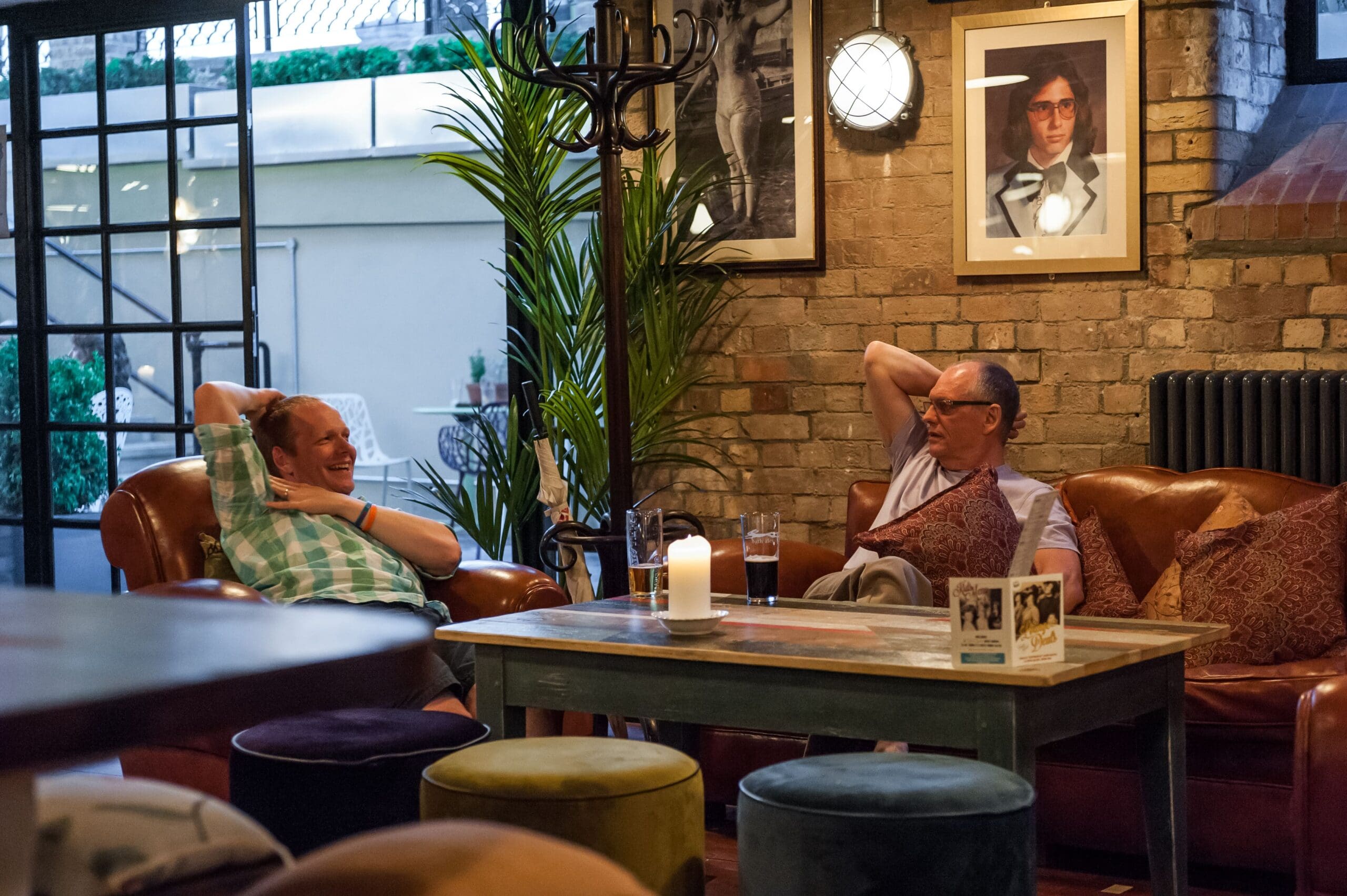 Two men relax and chat on leather sofas in a cozy, warmly lit Vivo lounge. Drinks and a candle sit on the table between them. Decor includes framed photos, plants, and brick walls with industrial-style windows in the background.