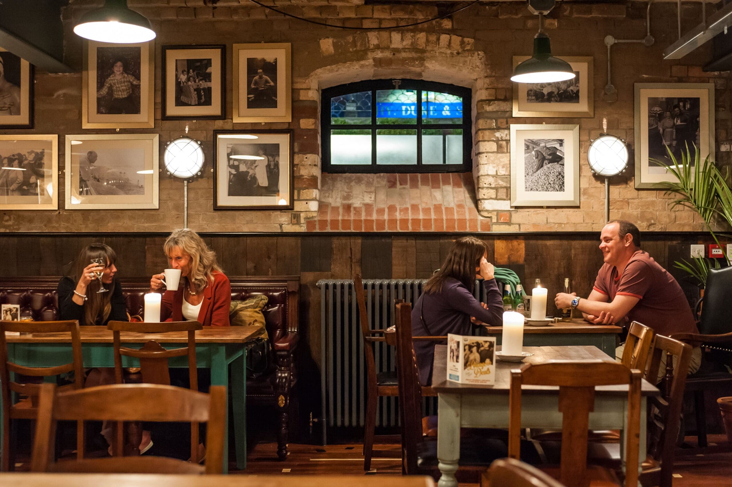 Two pairs of people sit at wooden tables in a cozy, warmly lit café with exposed brick walls, framed photos, and candles. The Vivo ambiance enhances their conversations as they enjoy drinks together.