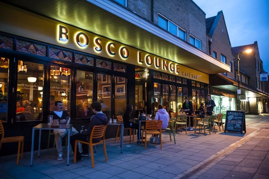 People sit at outdoor tables in front of Bosco Lounge café bar at dusk, chatting and dining under the warm glow of Bosco signage. The street is quiet, and the atmosphere appears relaxed yet lively.