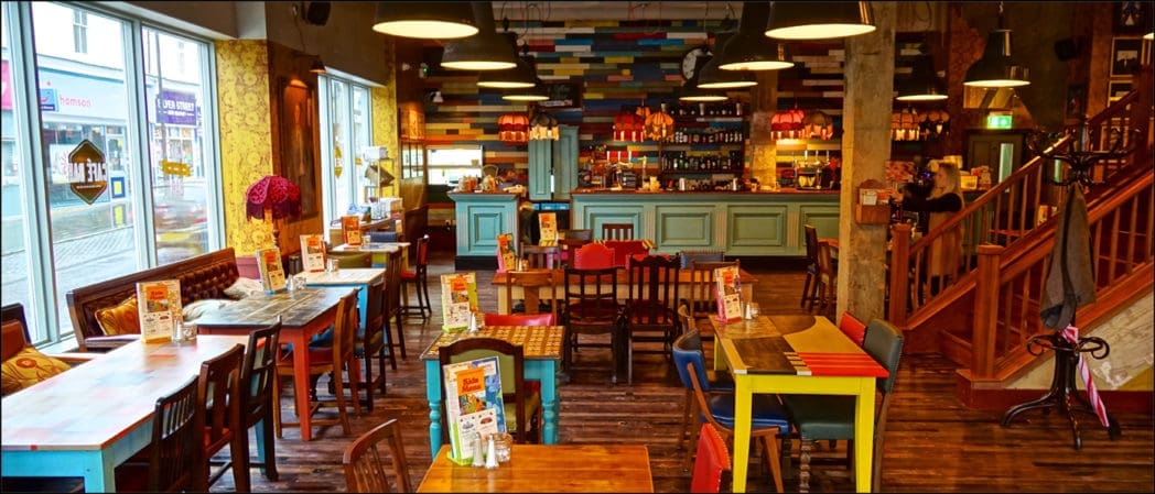 A colorful, eclectic café interior with mismatched tables and chairs, wooden floors, a teal Castello bar counter, vibrant wall decor, and large windows letting in natural light. Menus are placed on each table.