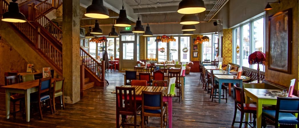 A brightly lit, cozy restaurant with colorful mismatched chairs and wooden tables, large windows letting in sunlight, hanging black pendant lights, and a staircase on the left side evokes the welcoming charm of Castello.