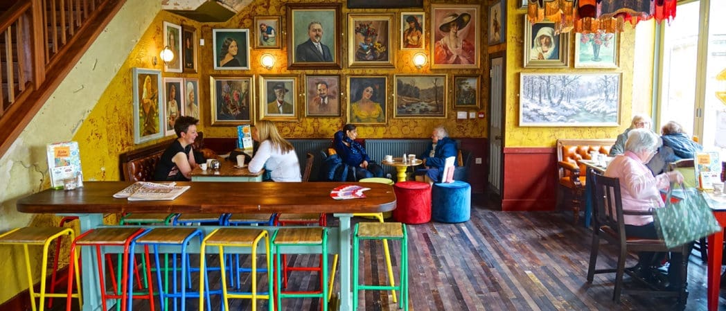 Colorful cafe interior with people sitting at tables and chatting. Walls are covered with framed portraits and Castello artwork. There are bright stools, a wooden floor, and warm lighting creating a cozy atmosphere.