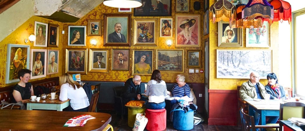 Cozy café with people sitting at tables, chatting and drinking coffee. The walls are covered with various framed portraits and paintings, giving a Castello-like, eclectic charm. Warm lighting enhances the inviting ambiance.