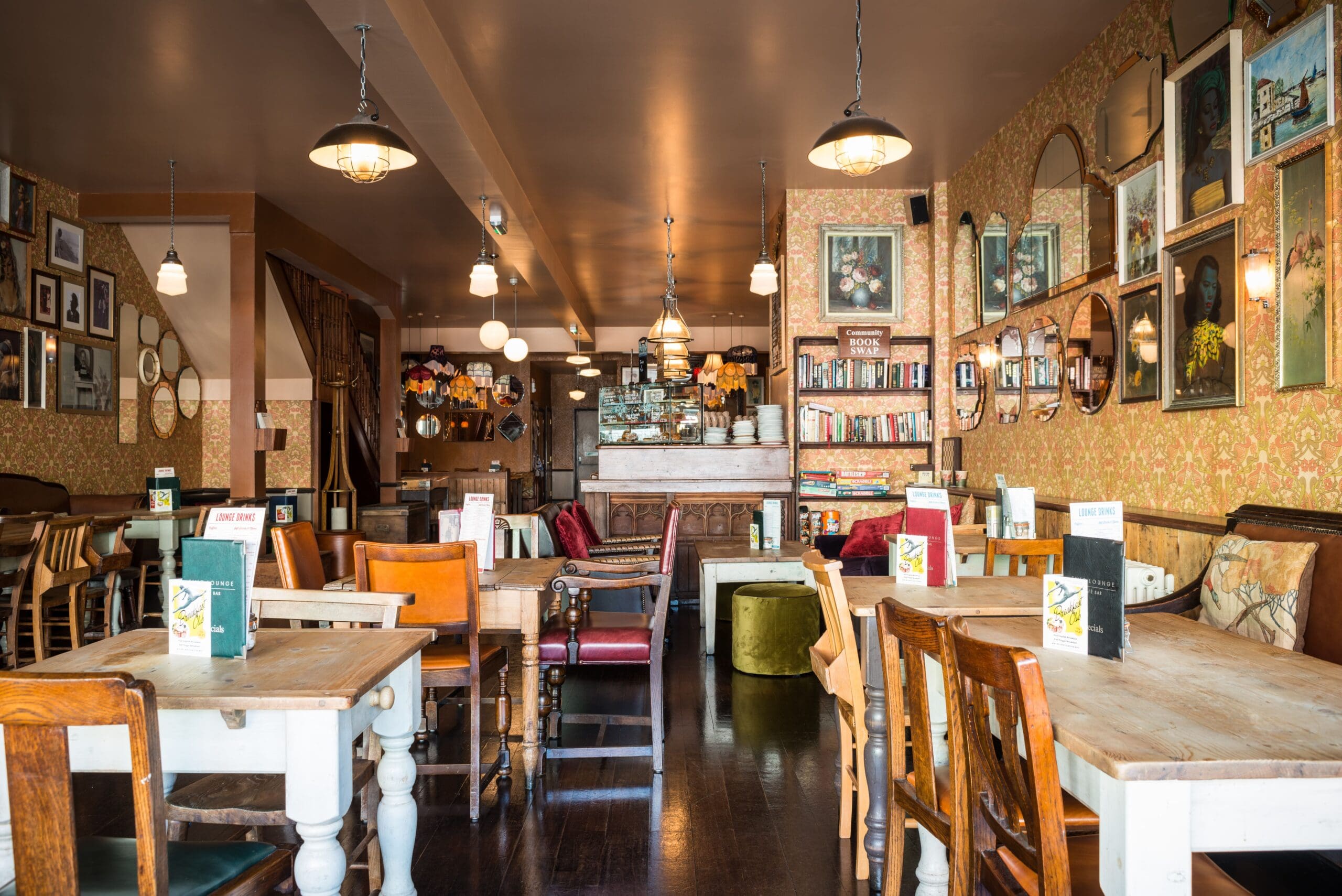 Eclectic vintage-style café interior with mismatched wooden tables and chairs, patterned wallpaper, and Fino touches throughout. Framed pictures, mirrors, and bookshelves add charm, while warm lighting creates a cozy atmosphere. Menus rest on each table.