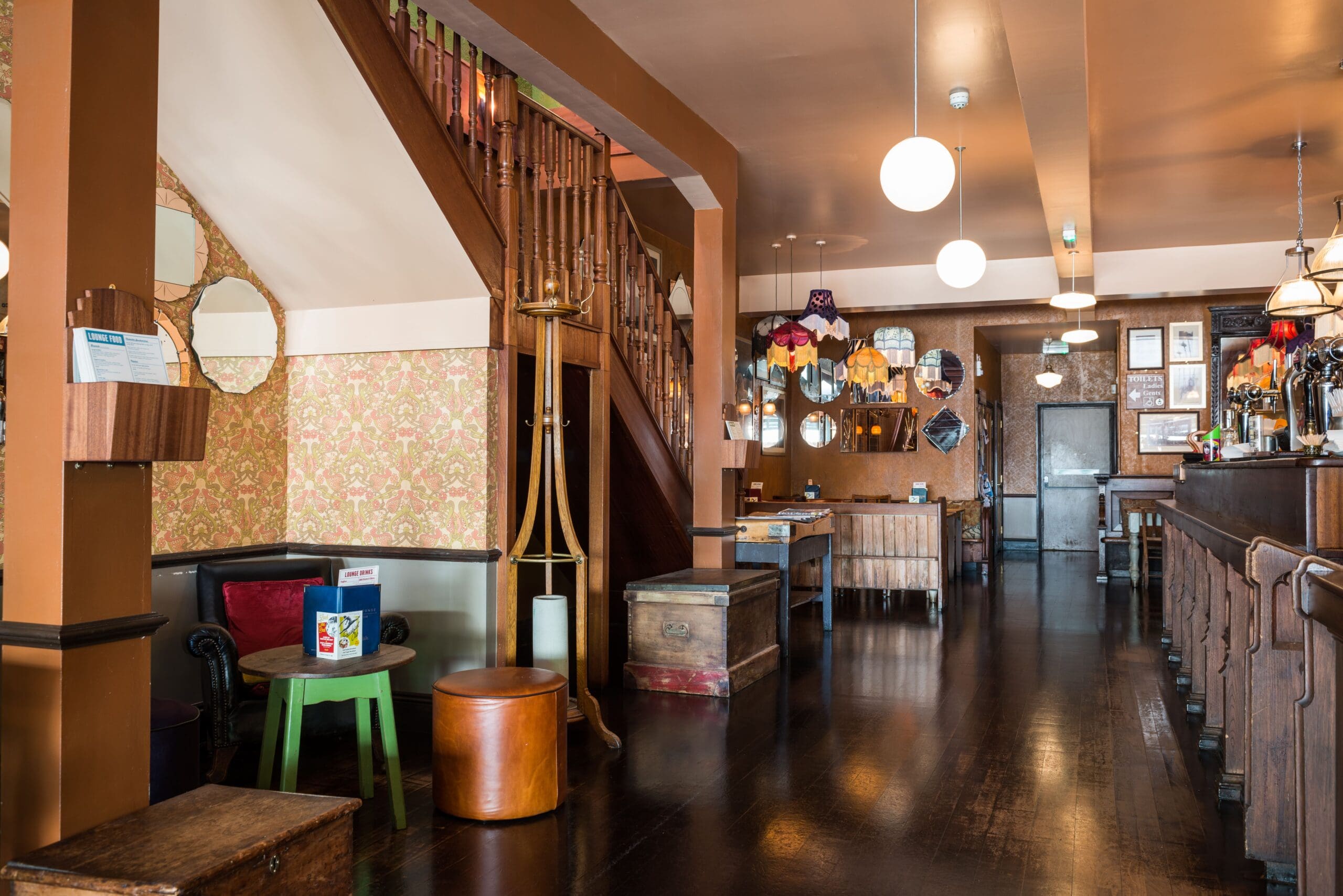 Cozy pub interior with wooden floors, a staircase, patterned wallpaper, Fino sherry on display, round hanging lights, vintage mirrors, eclectic furniture, and a bar on the right side. The space is warmly lit and inviting.