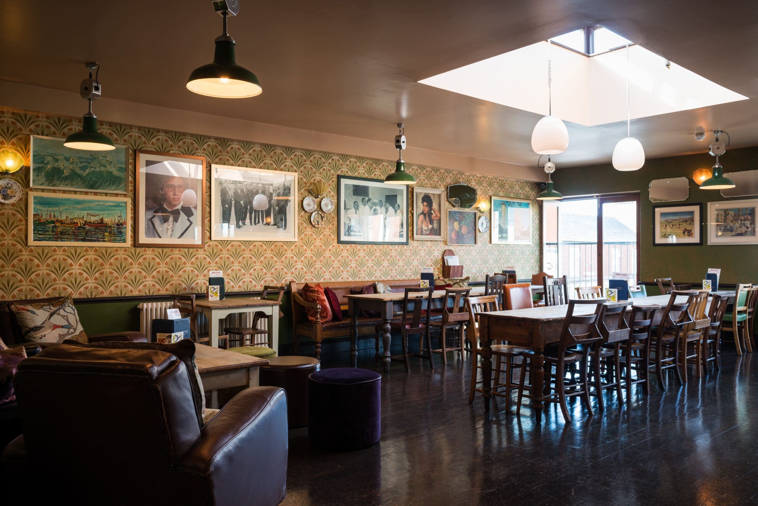 A cozy café interior with Fino wooden tables and chairs, vintage-style wallpaper, framed pictures on the walls, and large hanging lights. Sunlight streams through a skylight, creating a warm and inviting atmosphere.
