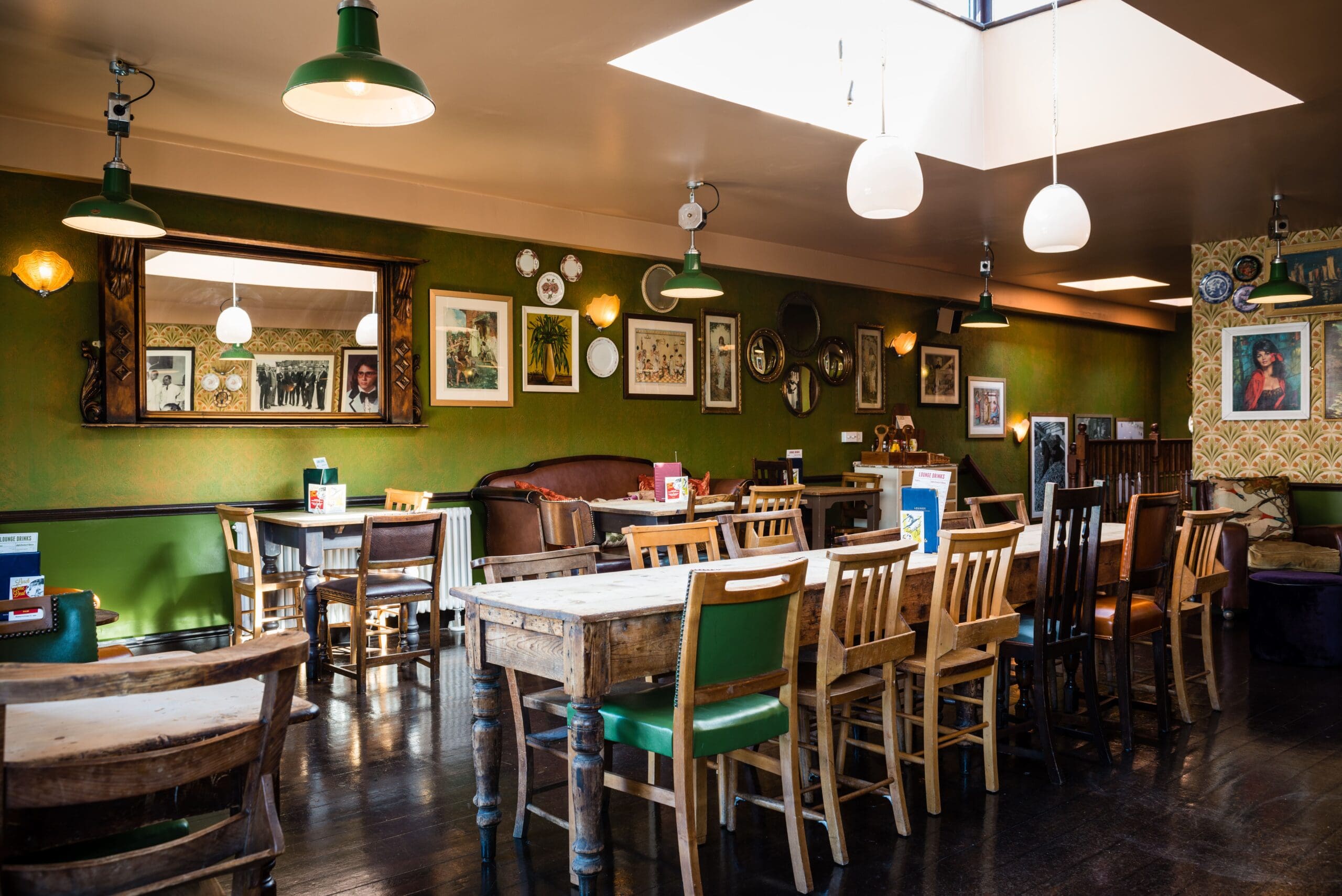 A cozy café with wooden tables and mismatched chairs, green walls decorated with framed pictures and mirrors, pendant lights hanging from the ceiling, and sunlight streaming through a Fino skylight.