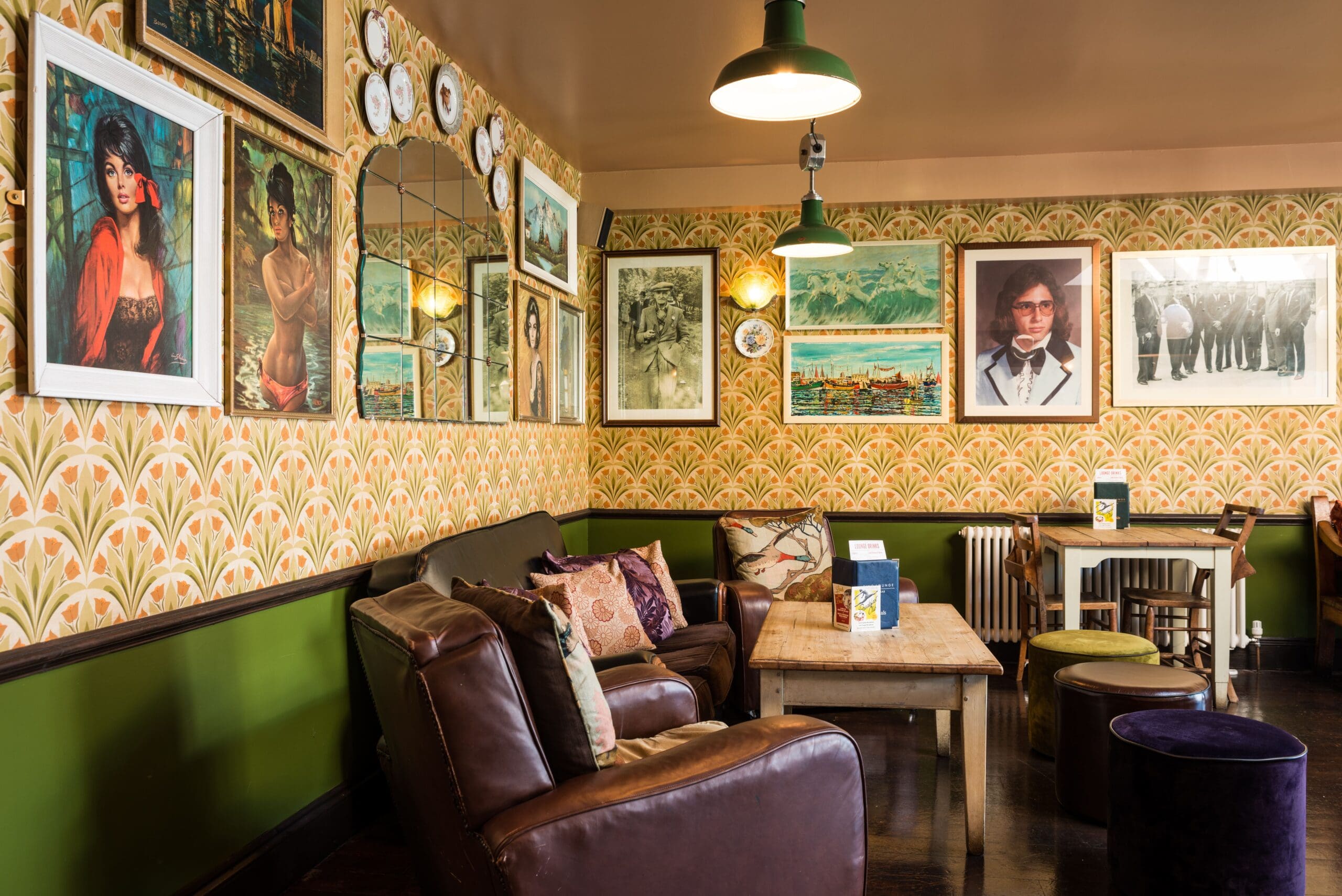A cozy, vintage-style lounge with Fino charm—brown leather chairs, wooden tables, patterned wallpaper, eclectic framed art, decorative plates, green hanging lights, and a mix of cushions and stools.