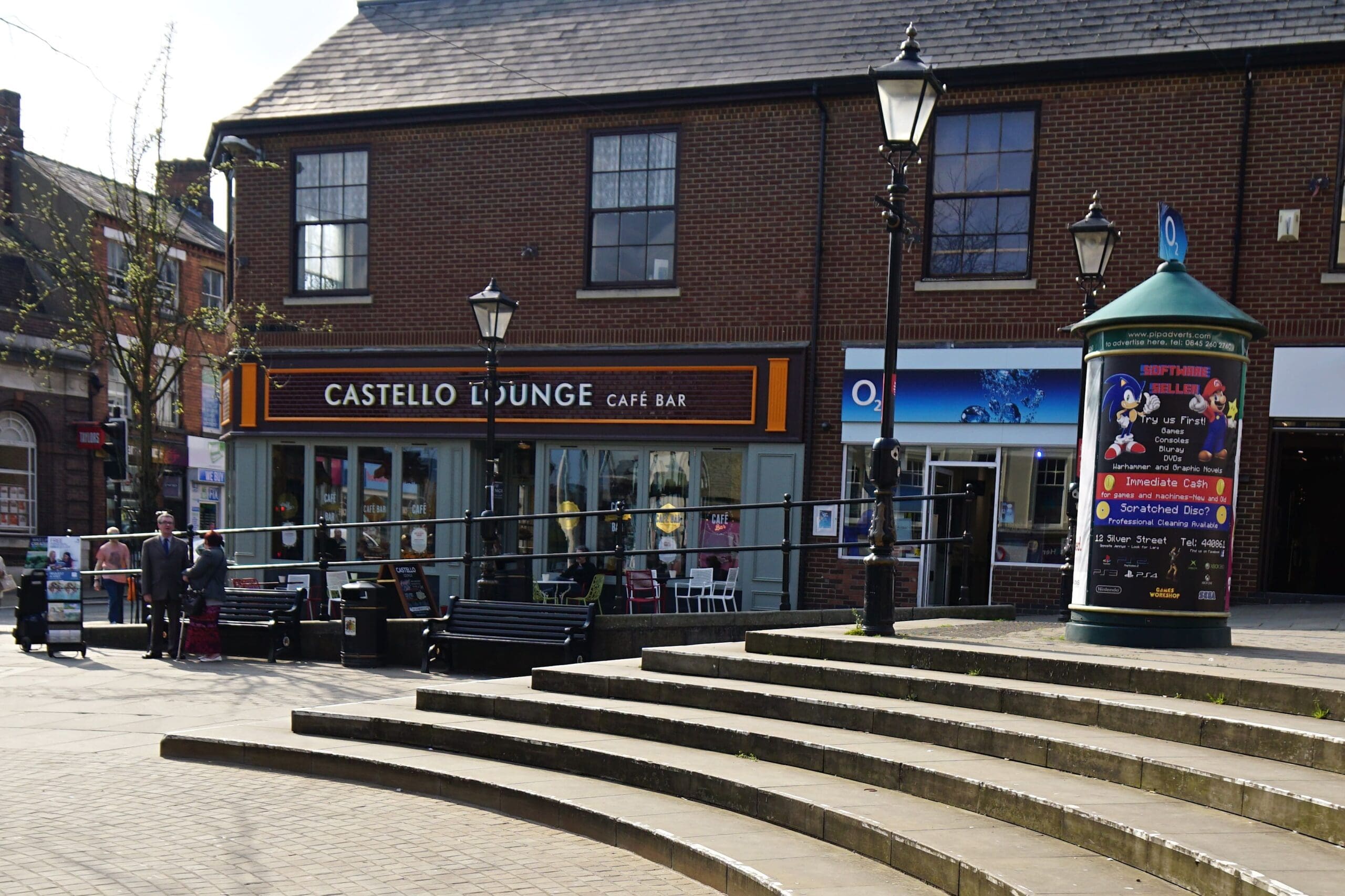Castello Lounge, a café bar with large windows, sits on a sunny street lined with benches, street lamps, and a cylindrical column covered in colorful posters. Steps lead up to the vibrant area in front of Castello’s shops.