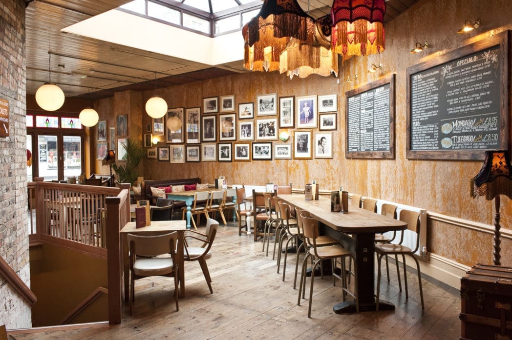 Cozy café Circo with wooden floors, vintage chairs, a long communal table, and eclectic lamps. Walls feature framed photos and chalkboard menus. Large windows and skylights fill Circo with natural light.