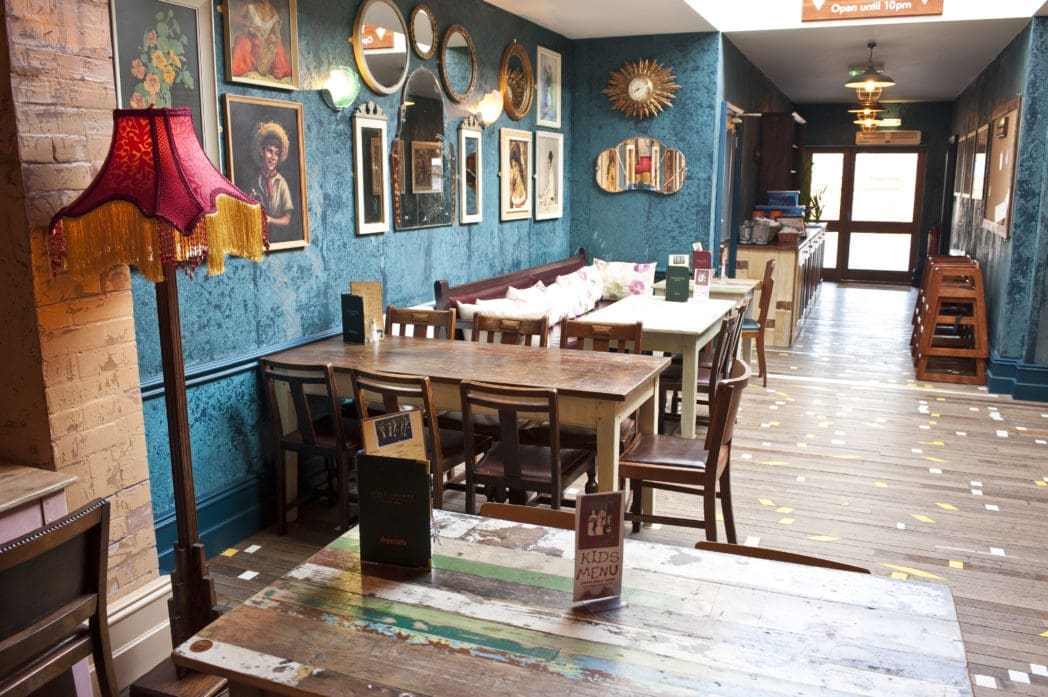 A cozy café interior with mismatched wooden tables and chairs, vintage lamps, teal walls decorated with framed art and mirrors, and a long cushioned bench along one side. Warm Circo lighting creates an inviting atmosphere.