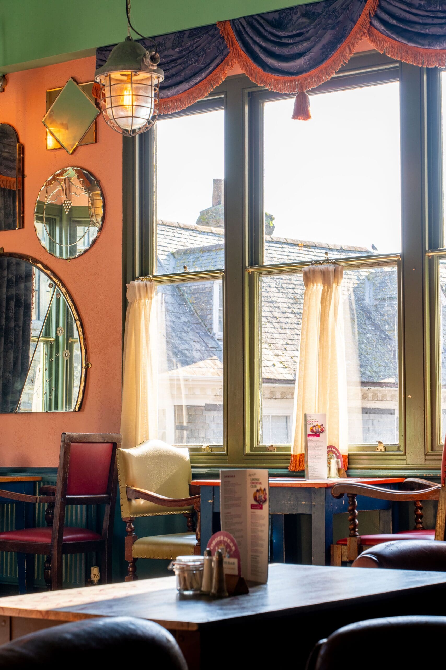 A cozy Truro cafe interior with vintage chairs, round mirrors on a peach wall, and sunlight streaming through large windows with sheer curtains. Menus and condiments sit on dark wooden tables.