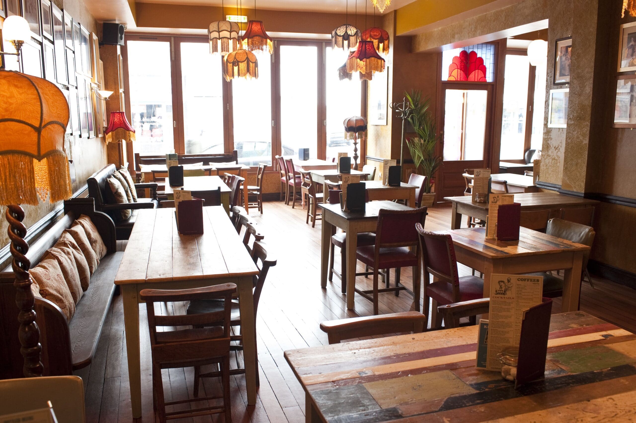 A cozy, sunlit café with wooden tables, mismatched chairs, vintage lamps, and large windows letting in natural light. Menus and chilled bottles of Modelo sit on the tables, adding to the warm and inviting atmosphere.
