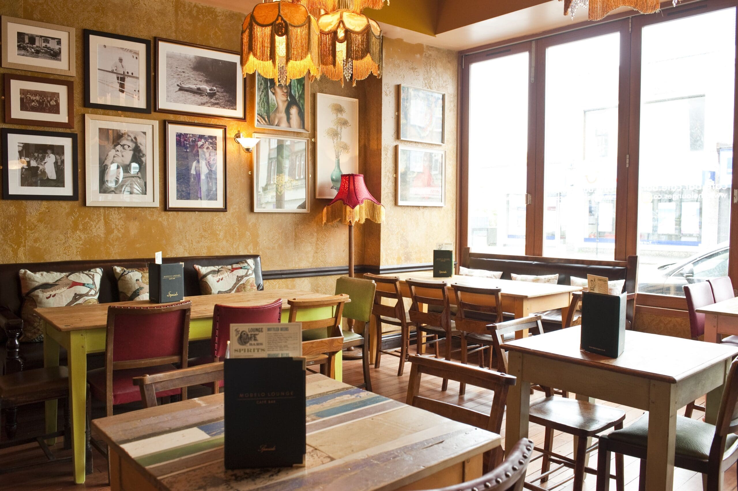A cozy café interior with wooden tables and mismatched chairs, vintage lamps, framed photos on textured walls, and daylight streaming through large windows. A cold Modelo sits beside the menus placed on each table.