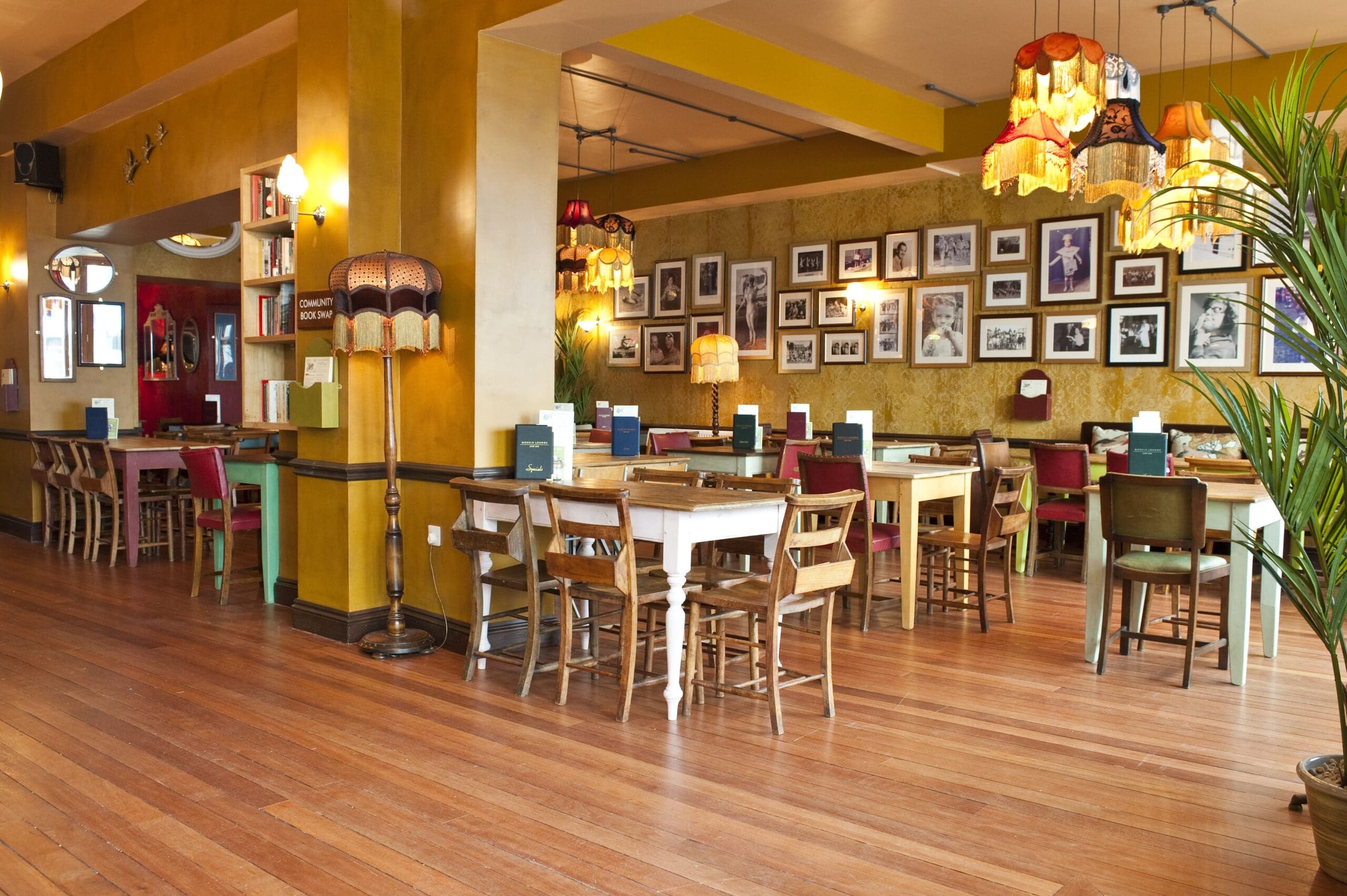 A cozy café with wooden floors, mismatched tables and chairs, vintage lamps, and a gallery wall of framed black-and-white photos—perfect for sipping a Modelo and soaking in the warm, eclectic atmosphere.
