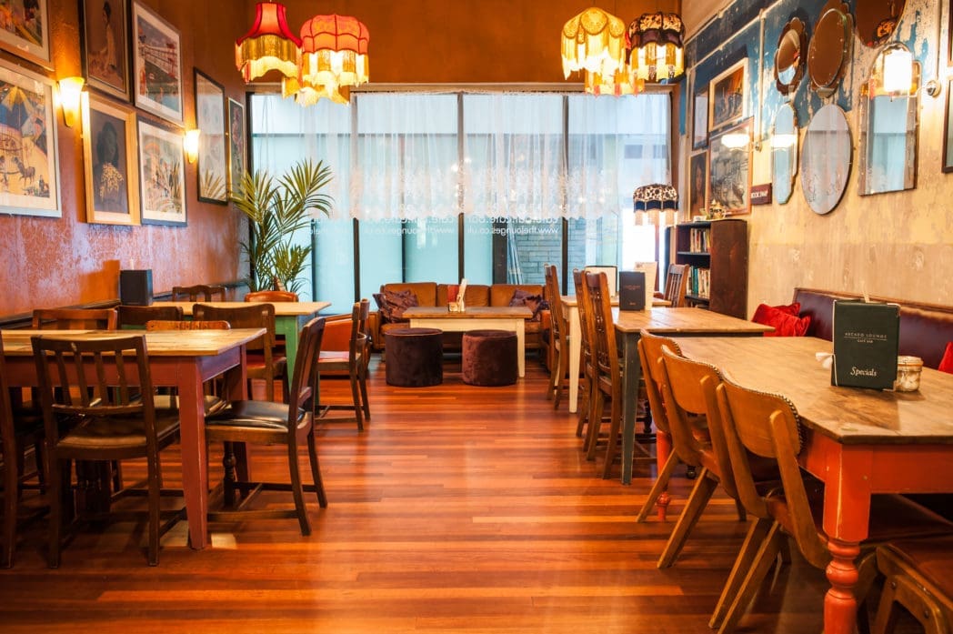 A cozy café with Arcado wooden floors, vintage furniture, eclectic lamps, framed art on the walls, and natural light coming through sheer curtains by a sofa in the back.