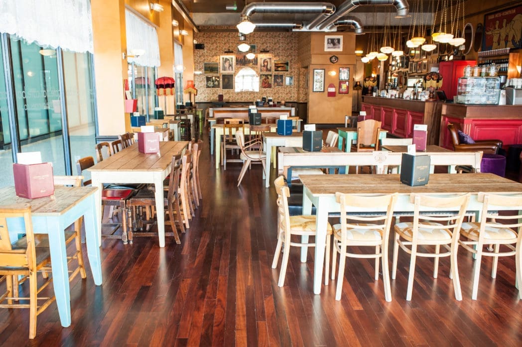 A cozy, vintage-style café with wooden tables and mismatched chairs, menus placed on each table, warm lighting, and retro Arcado decor. The space features polished wooden floors and a red counter at the back.
