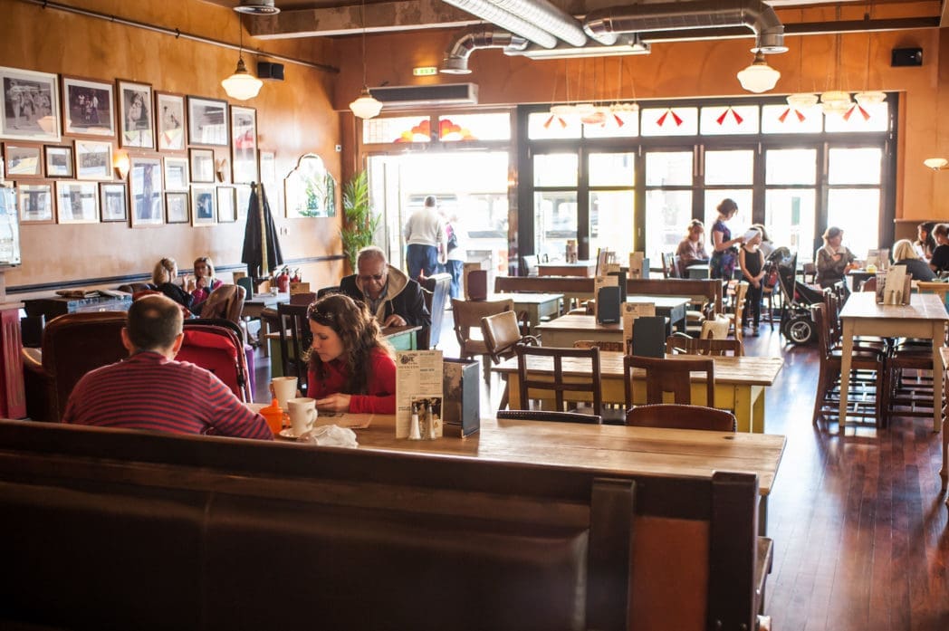 A cozy café with Arcado wooden tables and chairs, several groups of people sitting and talking, large windows letting in natural light, and framed photos hanging on the orange walls.