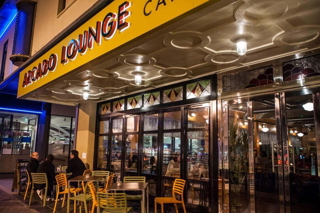 Outdoor seating at Arcano Lounge Café at night, where people sit at tables under a yellow Arcado sign. The interior is brightly lit and visible through large glass windows and doors.