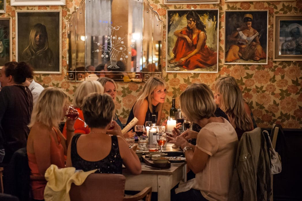 A group of women sit around a table in Arcado, a cozy, dimly lit restaurant, enjoying food and drinks. The floral wallpaper, framed portraits, candles, and wine bottles create a warm and inviting atmosphere.