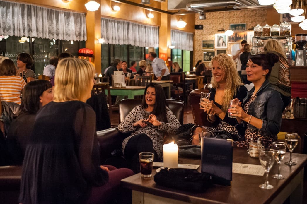 A group of women sit around a table in Arcado, a cozy, warmly-lit restaurant, laughing and talking. Drinks and a lit candle sit on the table as other patrons enjoy the lively atmosphere in the background.