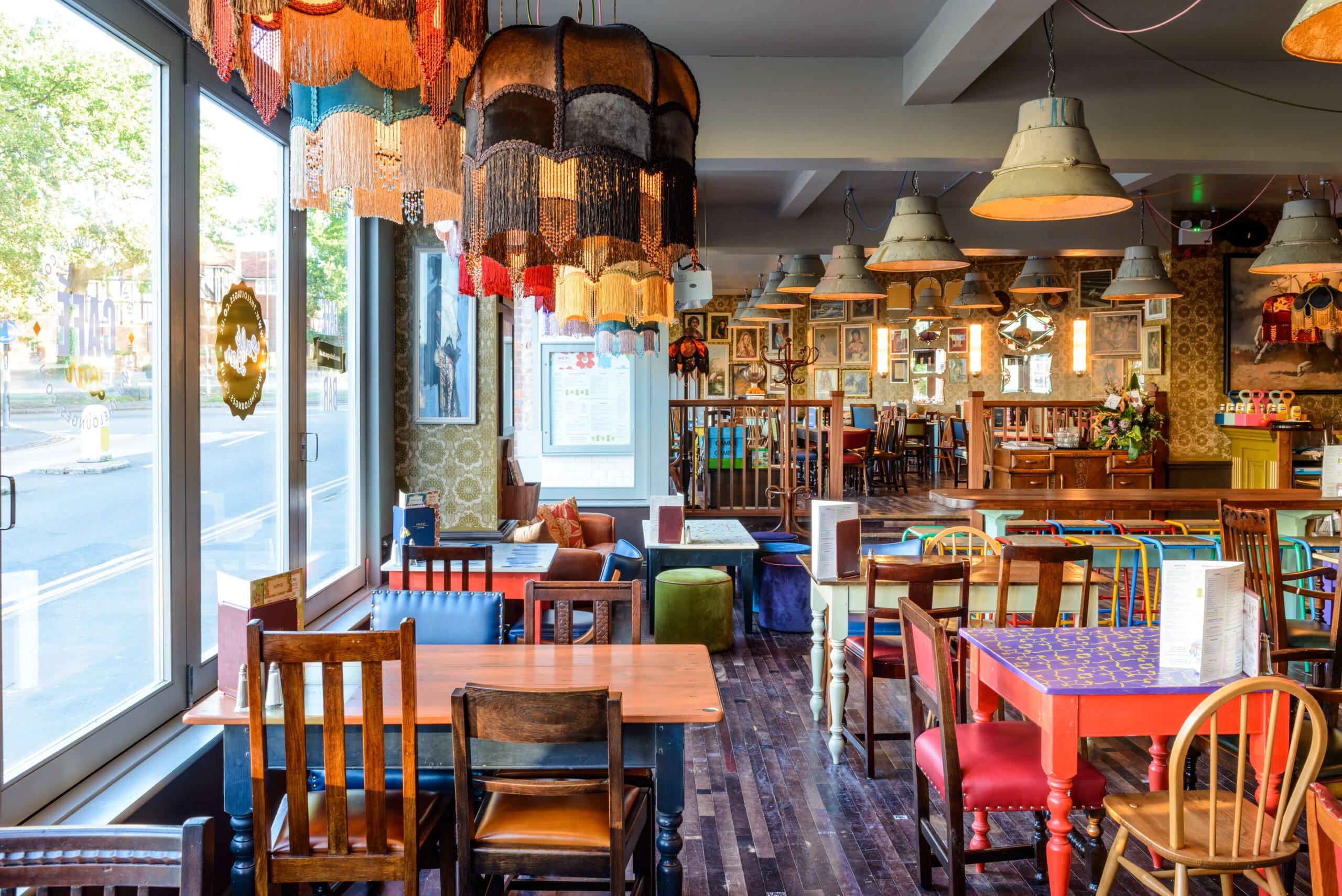 A colorful, eclectic cafe interior with mismatched tables and chairs, vintage lampshades, and framed art on patterned wallpapered walls gives off a vibrant Metro vibe. Sunlight streams in through large windows, brightening the inviting space.