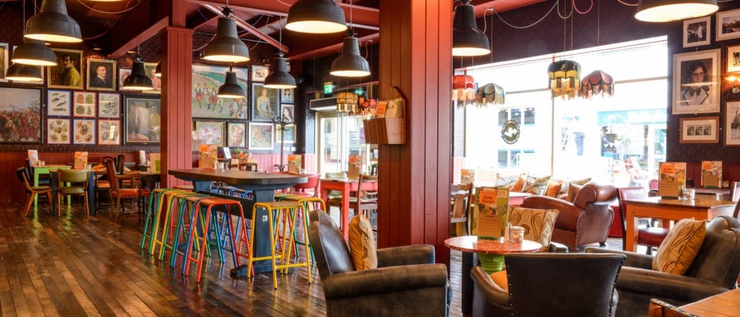 A cozy, eclectic café in Centro with colorful chairs, wooden tables, and armchairs. The walls are decorated with framed art and photos, while large windows let in natural light. Pendant lights hang from the ceiling.
