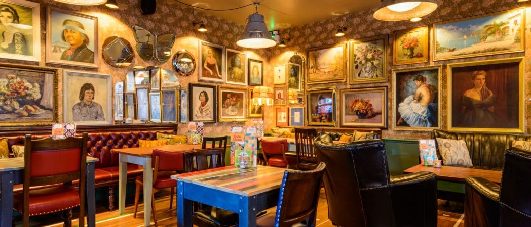 A cozy, eclectic café where "Croeso" (welcome) greets you, with colorful chairs and tables, decorated walls full of framed paintings and mirrors, warm lighting, and a relaxed, inviting atmosphere.