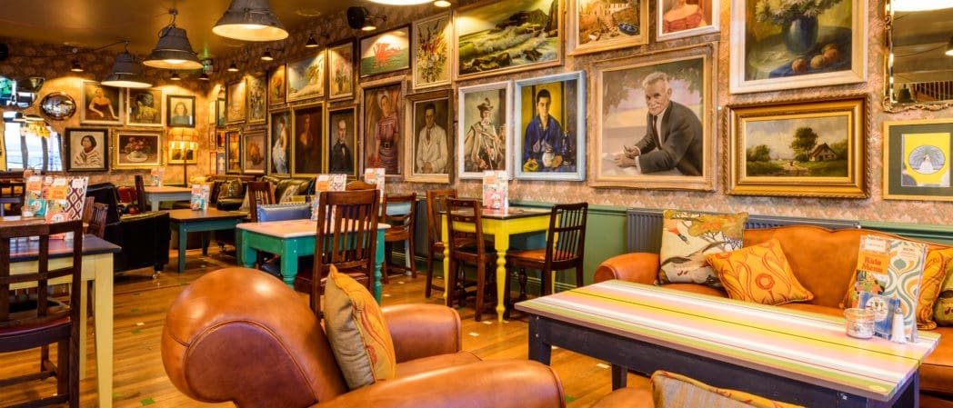 A cozy cafe with colorful mismatched chairs and sofas. The walls are covered with numerous framed paintings and portraits, creating an eclectic and inviting atmosphere. Warm lighting and a Croeso welcome enhance the vibrant decor.