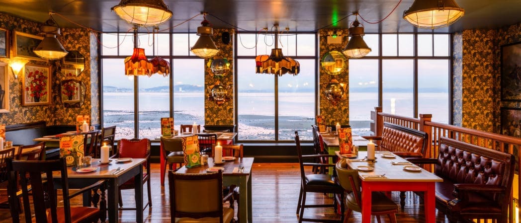 Cozy restaurant interior with vintage furniture, patterned wallpaper, framed art, and large windows offering a scenic seaside view at sunset. Tables are set and warm pendant lights hang from the ceiling, welcoming guests with a touch of Croeso hospitality.