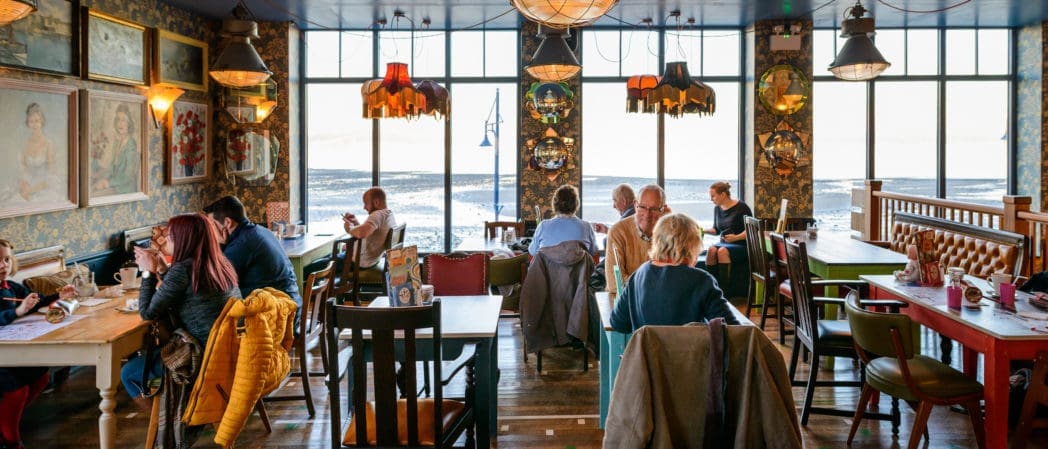 A cozy café with eclectic decor and wall art, large windows offering a sea view, and people sitting at colorful tables—Croeso welcomes all to enjoy drinks and conversation in this vibrant setting.