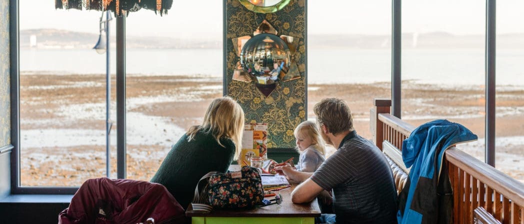 A family of three sits at a table by large windows overlooking a beach or bay. The adults and a young child are engaged in drawing or coloring together in a cozy Croeso restaurant or café.