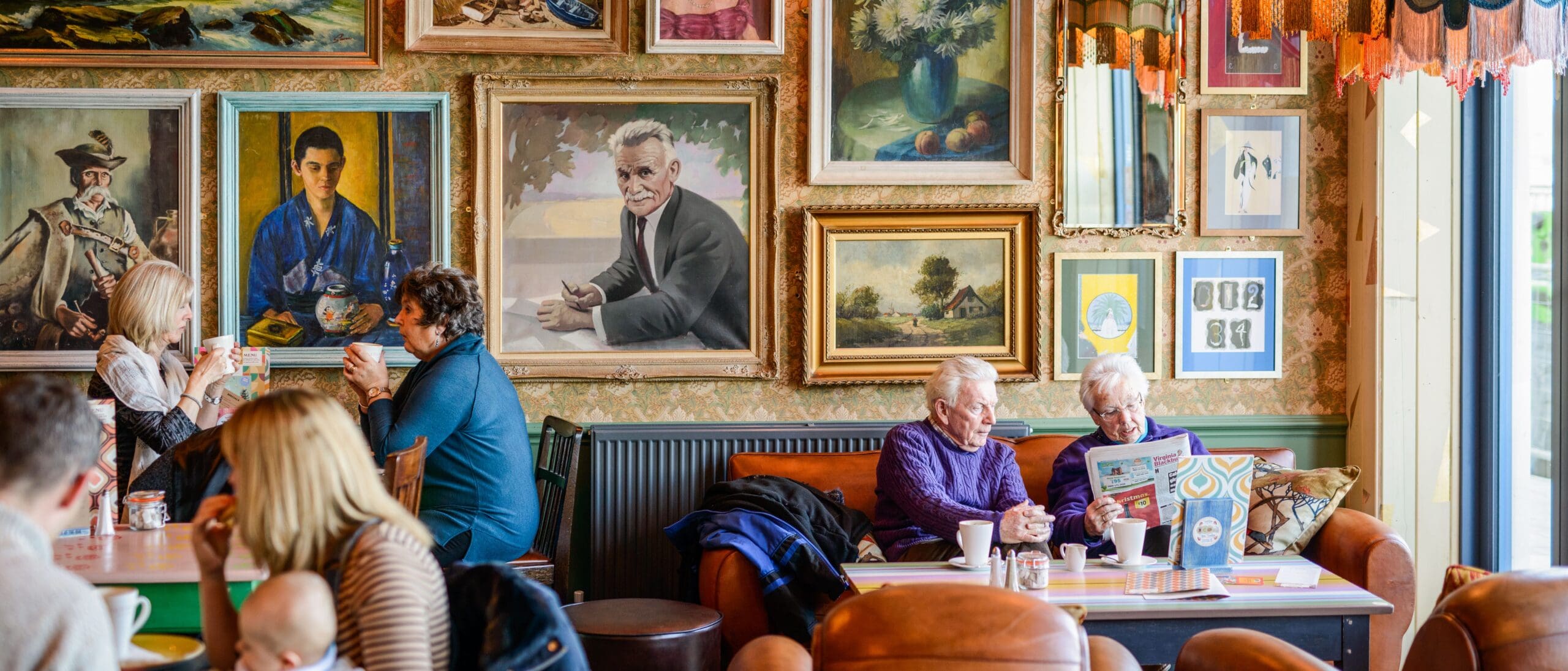 People sit and chat in a cozy cafe decorated with a gallery wall of colorful paintings and portraits. A “Croeso” sign welcomes guests as two elderly people read a newspaper on a sofa, while others enjoy drinks bathed in natural light from large windows.