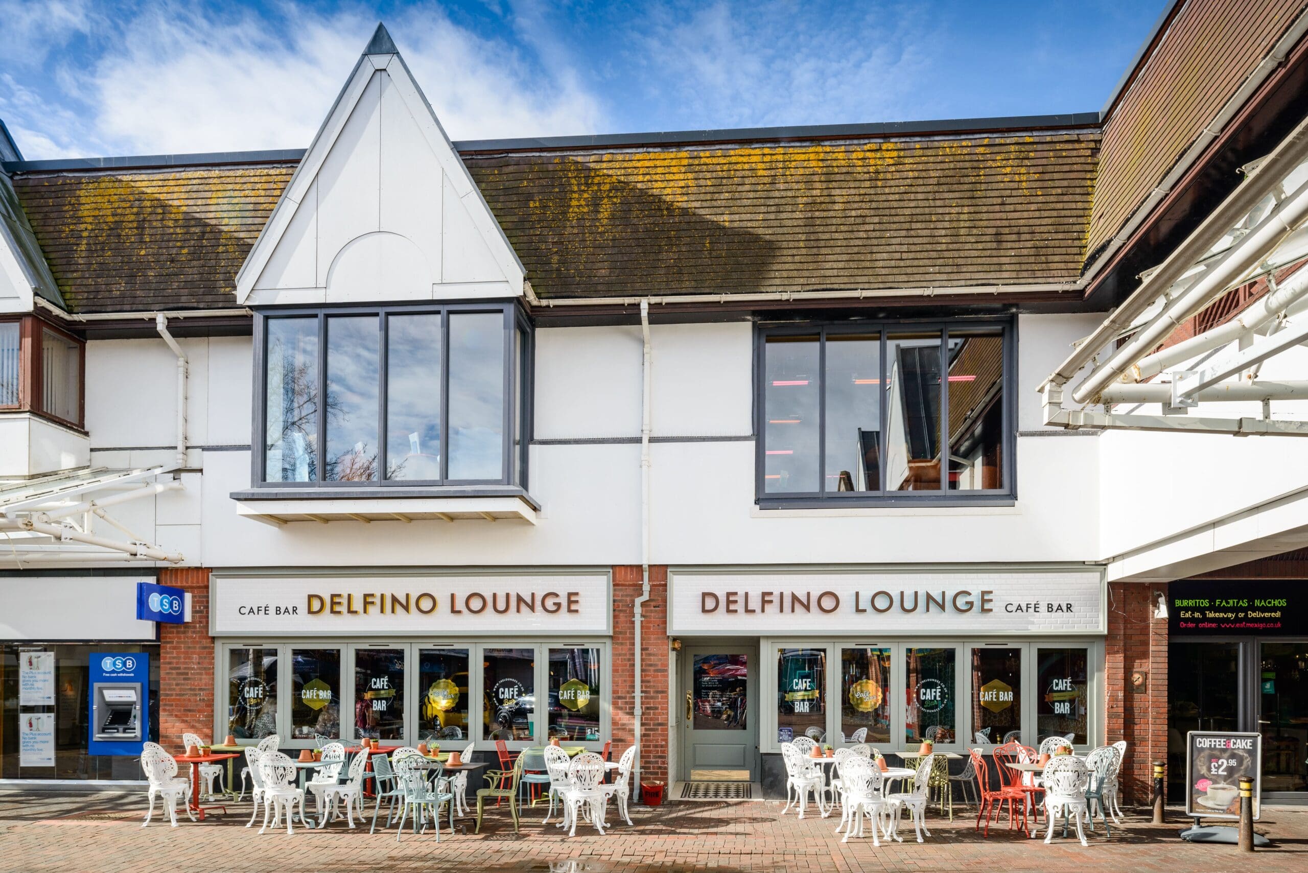 Outdoor seating area with white tables and chairs in front of Delfino Lounge café bar, located in a modern building with large Delfino signage and expansive windows under a clear blue sky.