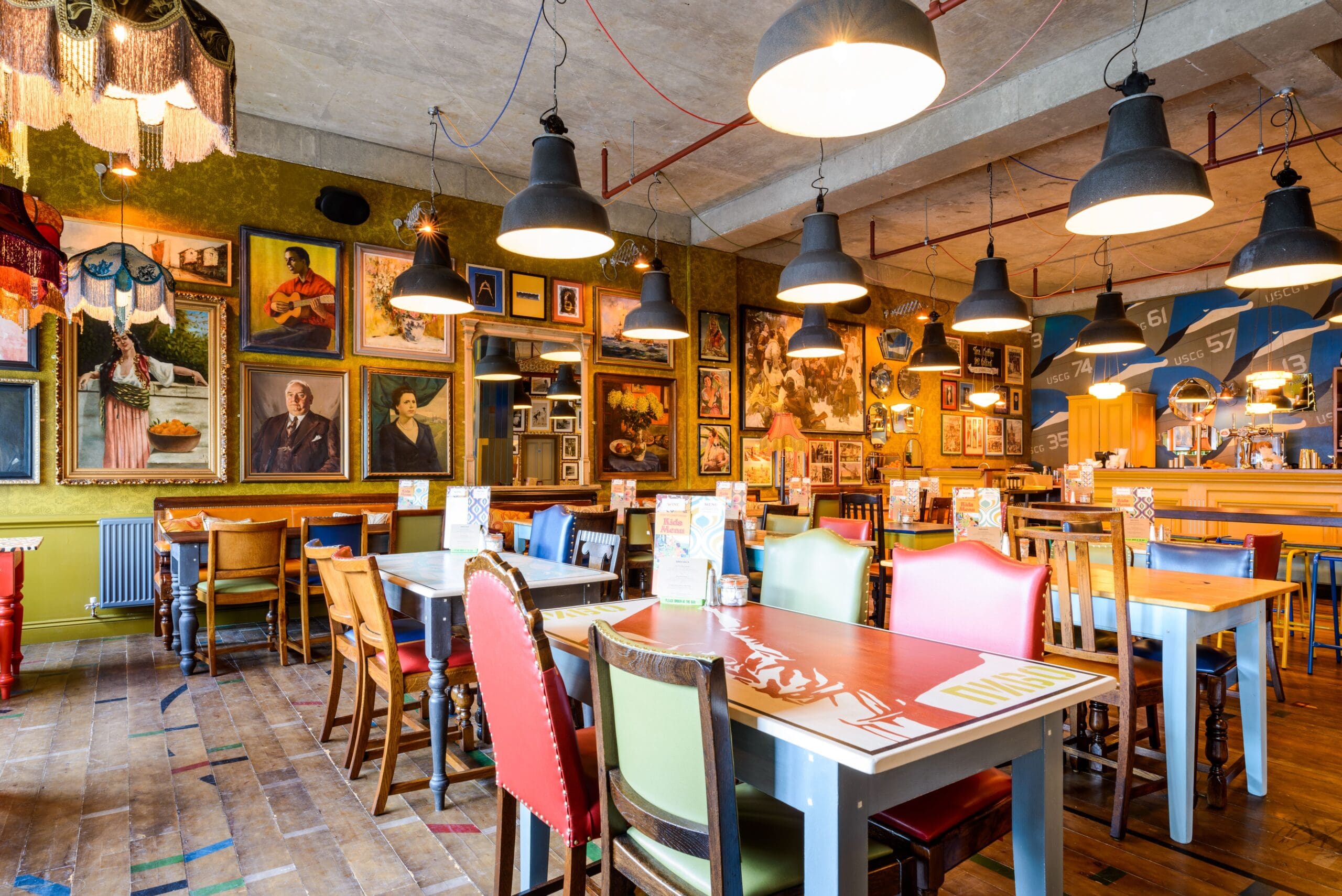 Colorful, eclectic restaurant interior with mismatched chairs and wooden tables. Delfino’s walls are decorated with numerous framed paintings and photos. Warm lighting hangs from the ceiling, creating a cozy, vibrant atmosphere.
