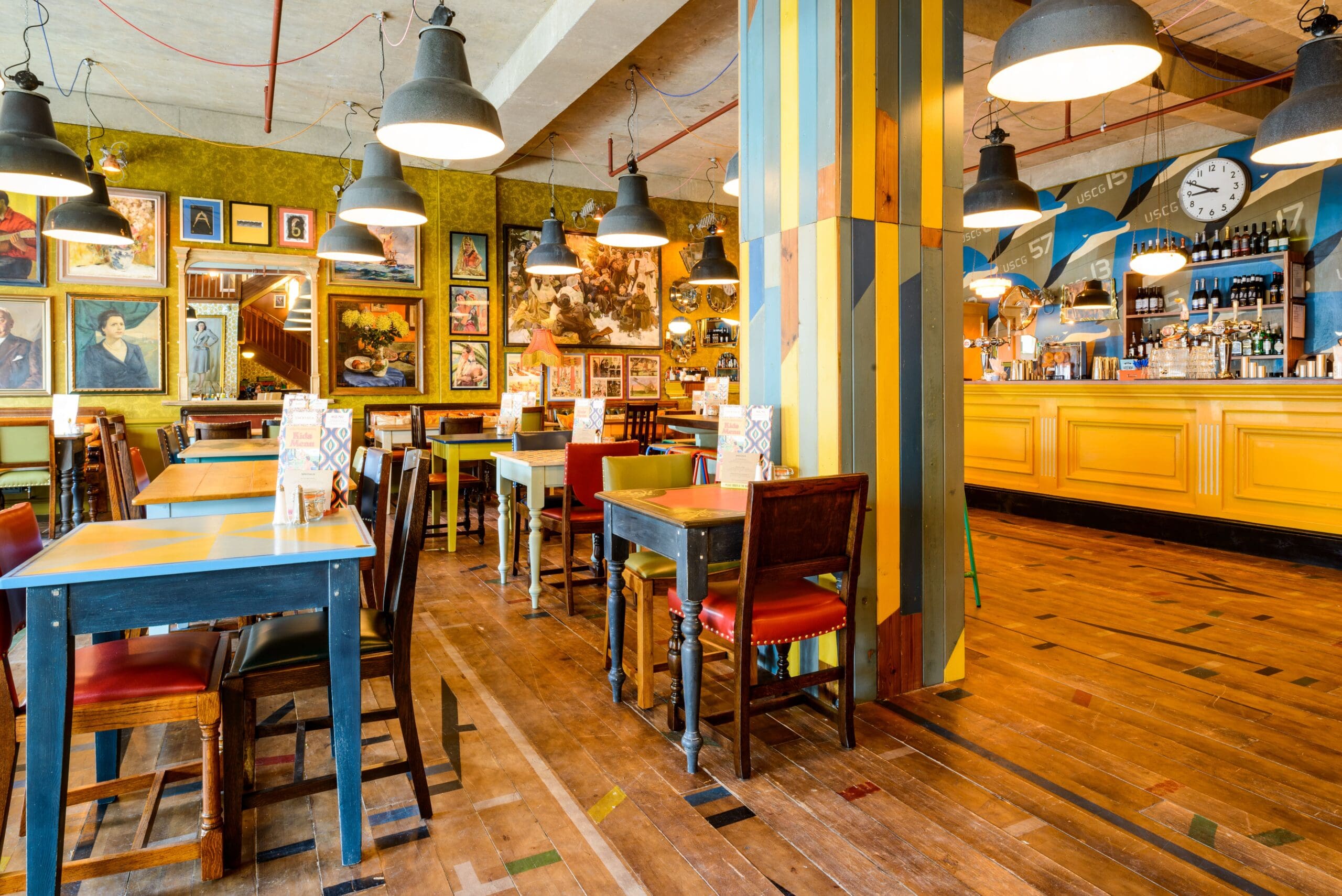 A brightly lit café with yellow and green décor, wooden tables and chairs, eclectic framed artwork on the walls, a striped pillar, and a Delfino-inspired bar area with shelves of bottles and a large clock.