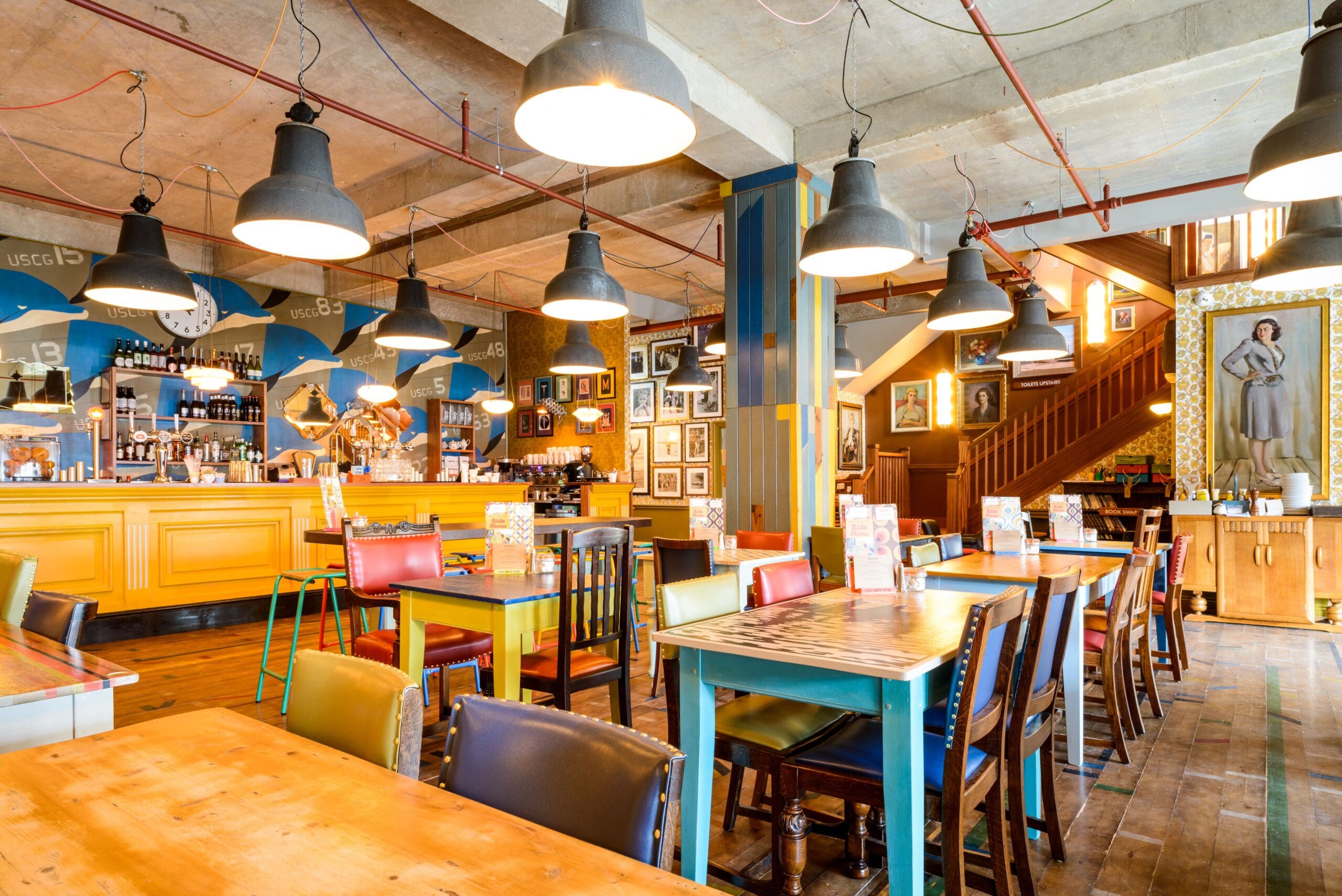 A brightly lit, eclectic café interior with colorful chairs, wooden tables, hanging lamps, Delfino art-covered walls, and a yellow bar counter. Stairs and framed portraits are visible in the background.