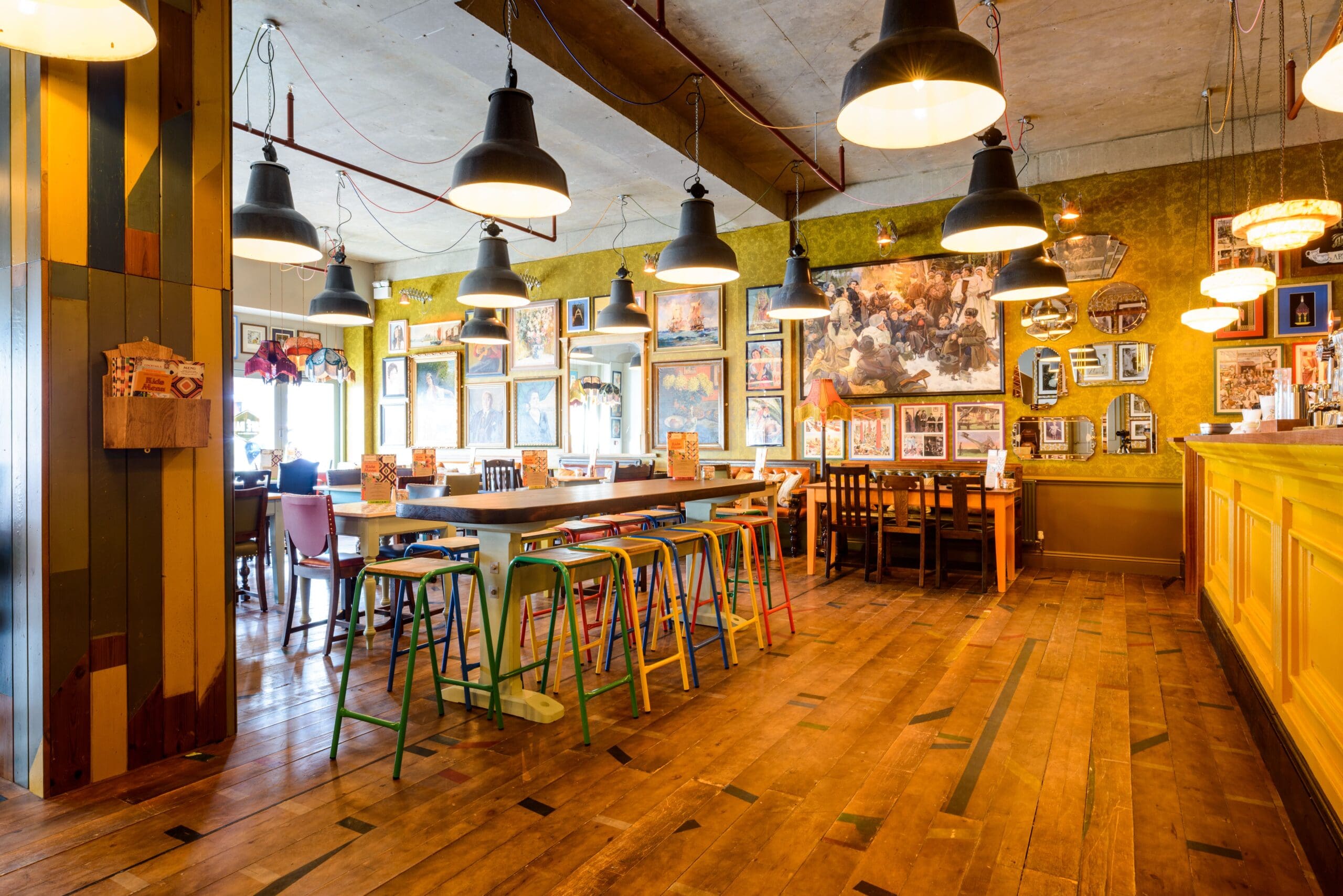 A cozy, eclectic restaurant interior with colorful chairs around wooden tables, pendant lights hanging from the ceiling, and walls decorated with various framed pictures and artworks. Delfino’s space has a warm, inviting atmosphere.