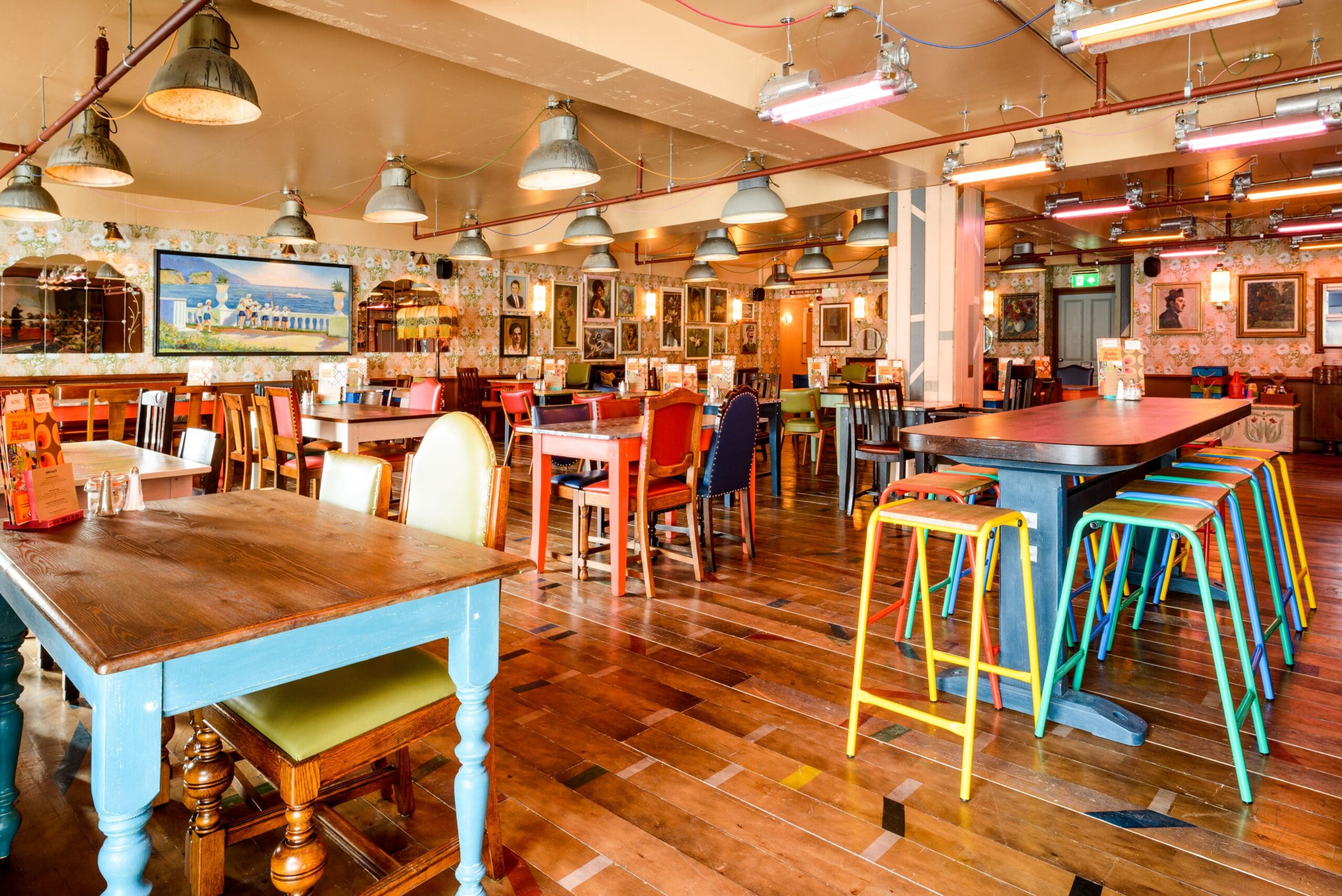 Colorful, eclectic Delfino restaurant interior with wooden floors, mismatched tables and chairs, vibrant bar stools, patterned wallpaper, and framed artwork covering the walls; pendant lights hang from the ceiling.